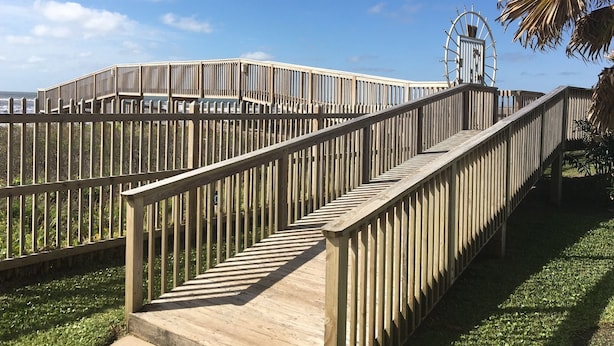 A wooden boardwalk with a spiral staircase leading to a viewing platform, surrounded by a fence and palm trees.