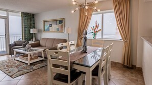 A modern living room with a glass coffee table, white chairs, a beige sofa, and a dining table with chairs. There is a chandelier, a painting on the wall, and a view through the window. - Direct Oceanfront 3BR/2BA, Park on same level as condo (Myrtle Beach)