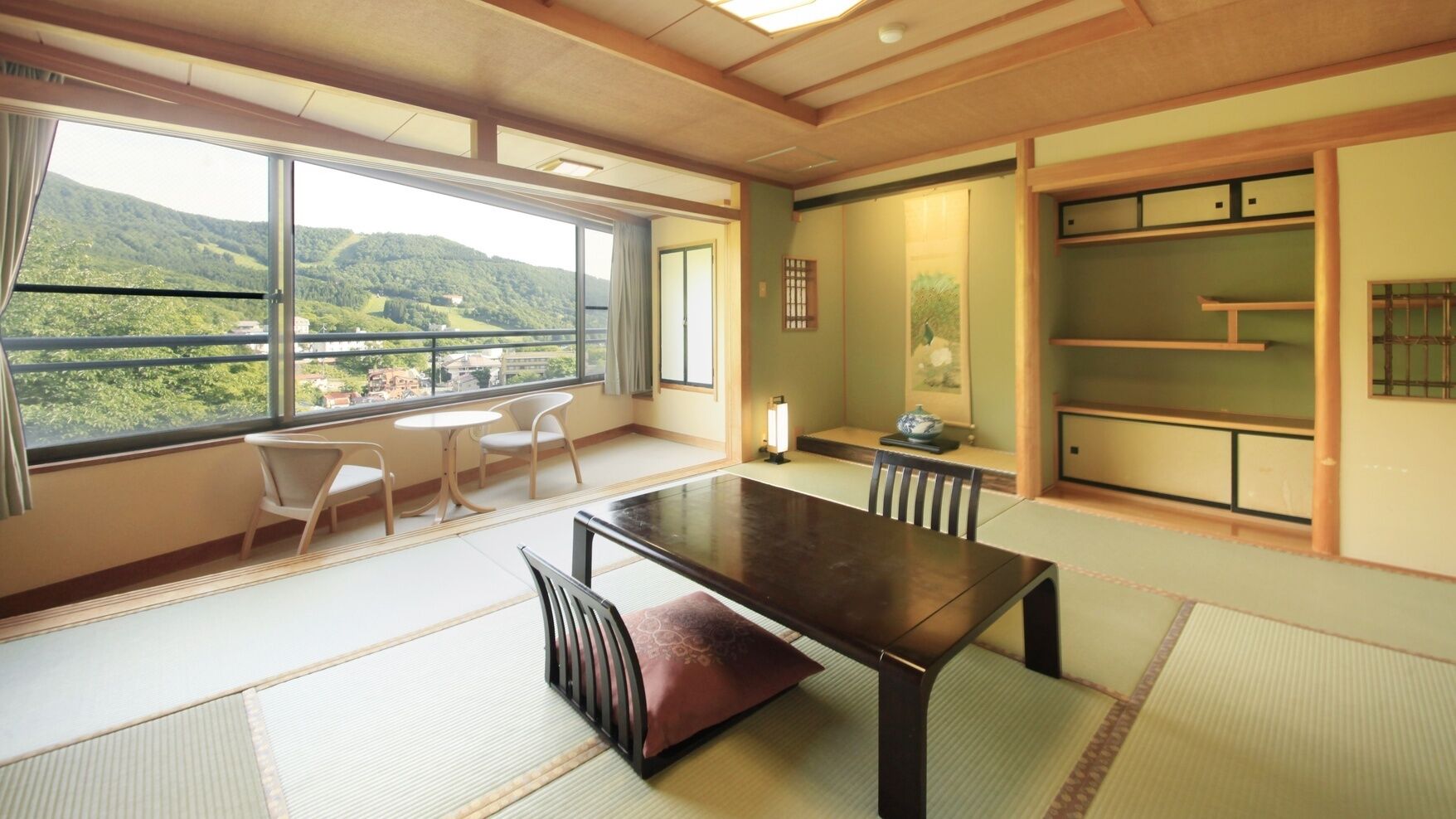 Quarto com piso de tatame, uma mesa baixa, cadeiras, vista para uma paisagem montanhosa através de grandes janelas e decoração tradicional japonesa.