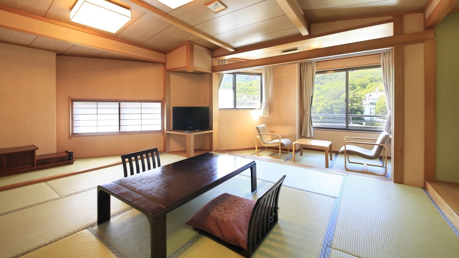 Um quarto japonês tradicional com tatames, uma mesa baixa, uma televisão e vista para uma paisagem verde através de grandes janelas.