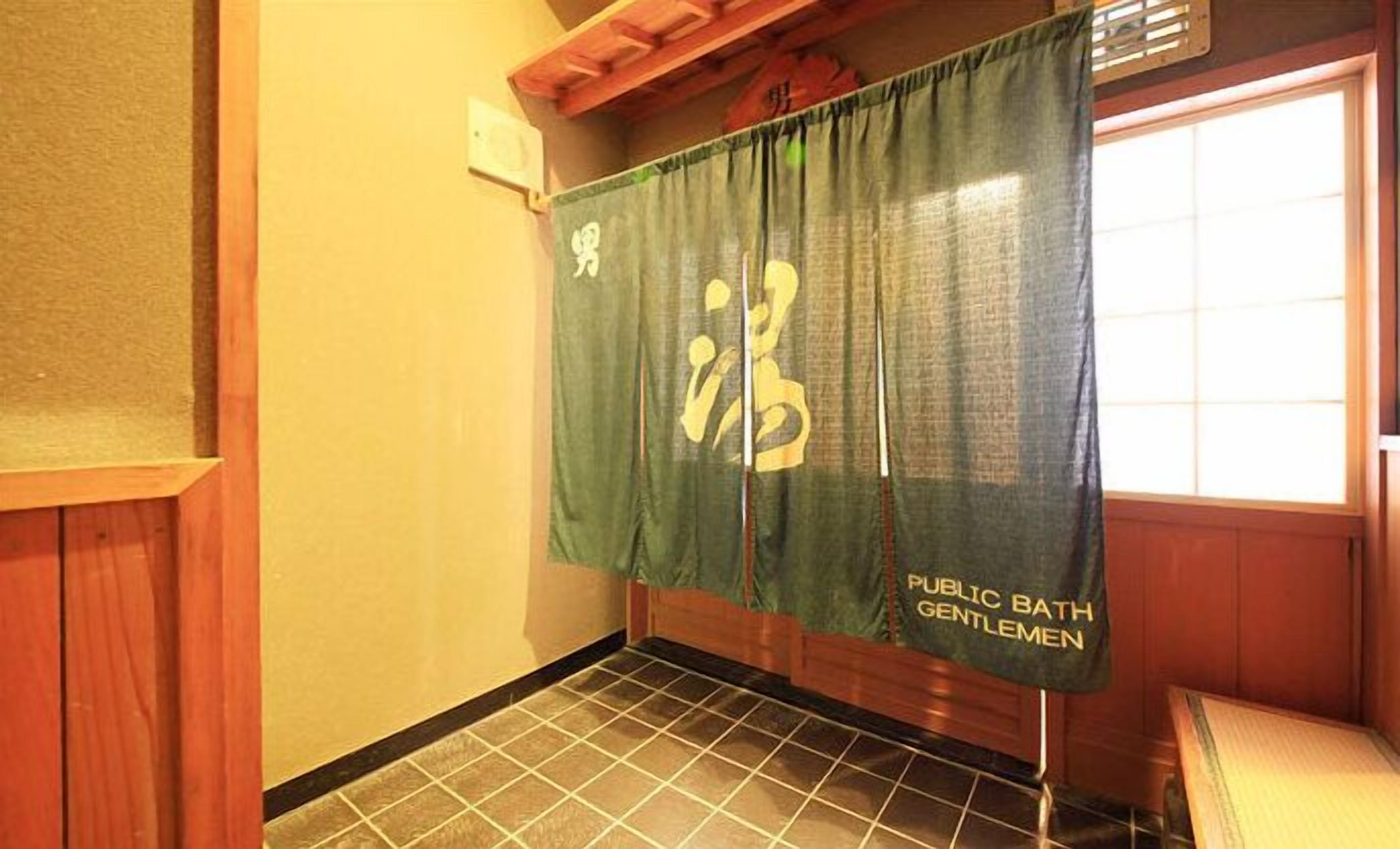 A bathroom with a tiled floor, a green curtain with Japanese characters, and a sign reading "Public Bath Gentlemen".