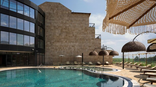 Un área de piscina moderna con una piscina azul cristalina, sillas de descanso y un edificio de piedra con grandes ventanales.