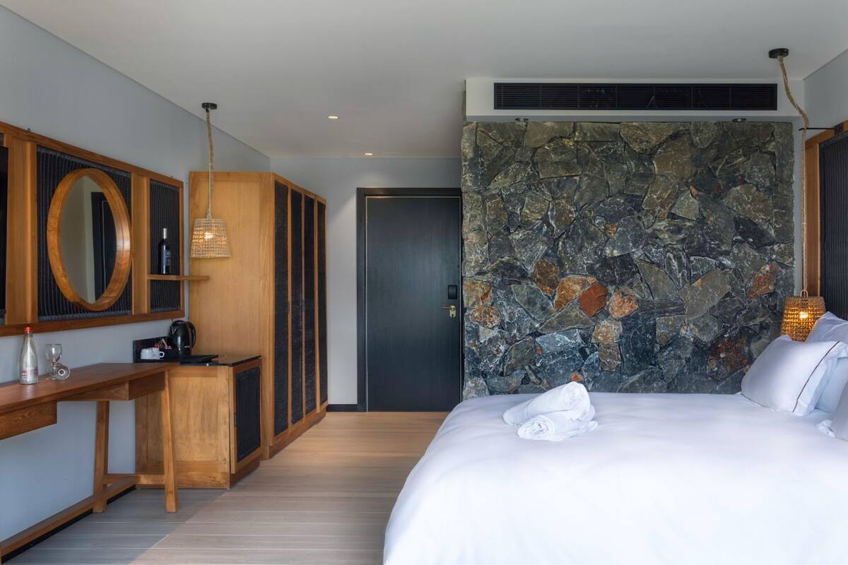A hotel room with a large bed, a wooden desk with a mirror, a stone wall, and a door.