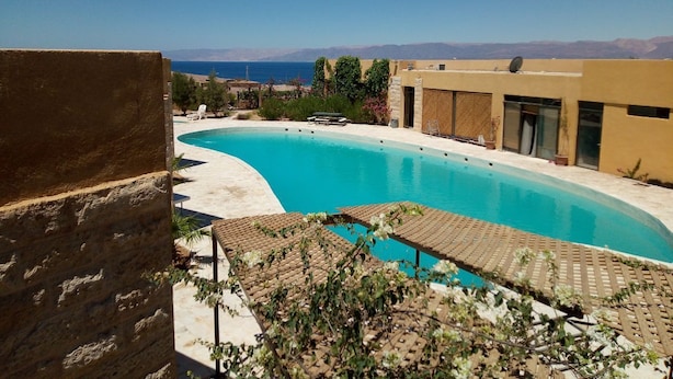 Une piscine à bord incurvé, entourée d’une terrasse aménagée avec des chaises longues, et un bâtiment à toit plat en arrière-plan.