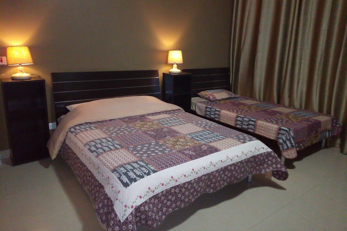 A hotel room with two beds, each with a quilted bedspread and a nightstand with a lamp.