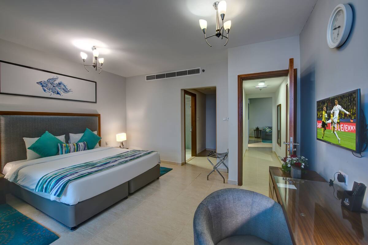 A modern hotel room with a large bed, a desk, a TV, and a clock on the wall.