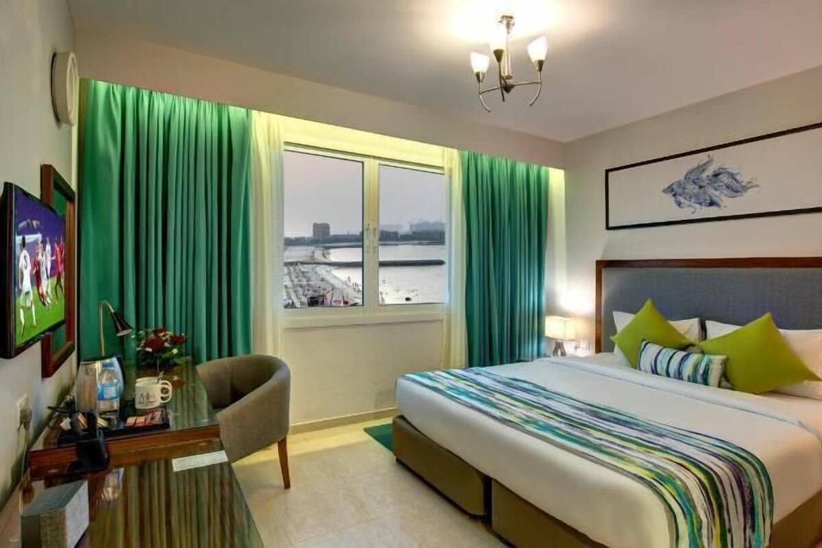 A hotel room with a bed, a desk, a chair, a window with curtains, a painting, and a view of a bridge.