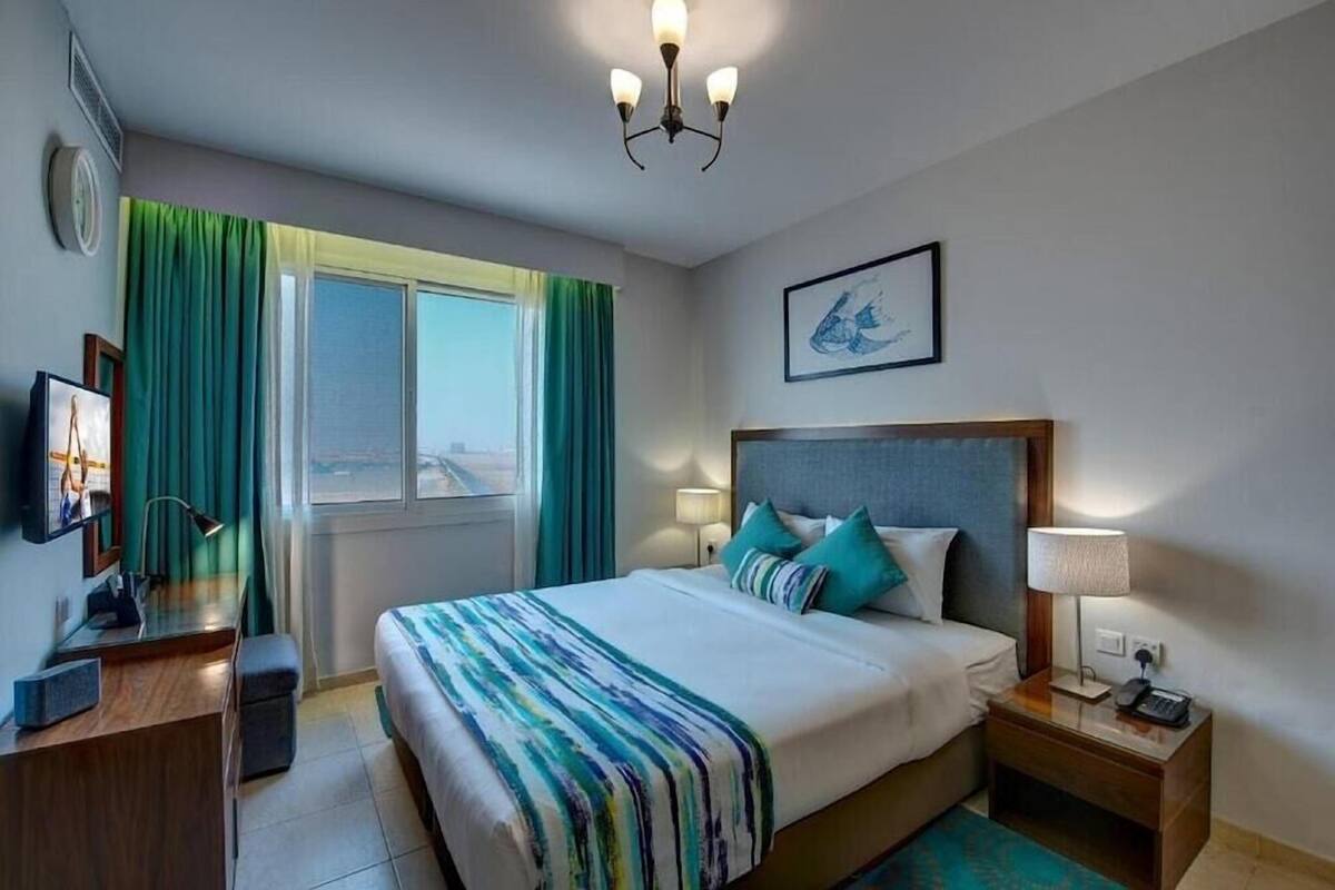 Una camera d'albergo con un letto, comodini, una scrivania, una sedia, una finestra con tende, un lampadario e un quadro appeso al muro.