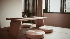 Una mesa de madera con una base de taburete tejido, ubicada frente a una ventana con persianas verticales.