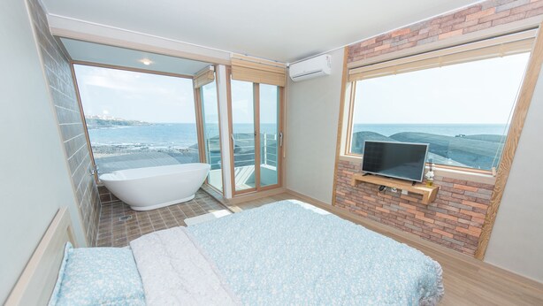 A modern bathroom with a bathtub, a large window with a view of the sea, and a TV on a wooden shelf. - Pension Jung Won (Seogwipo)