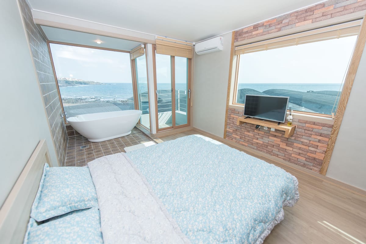 A modern bathroom with a bathtub, a large window with a view of the sea, and a TV on a wooden shelf.