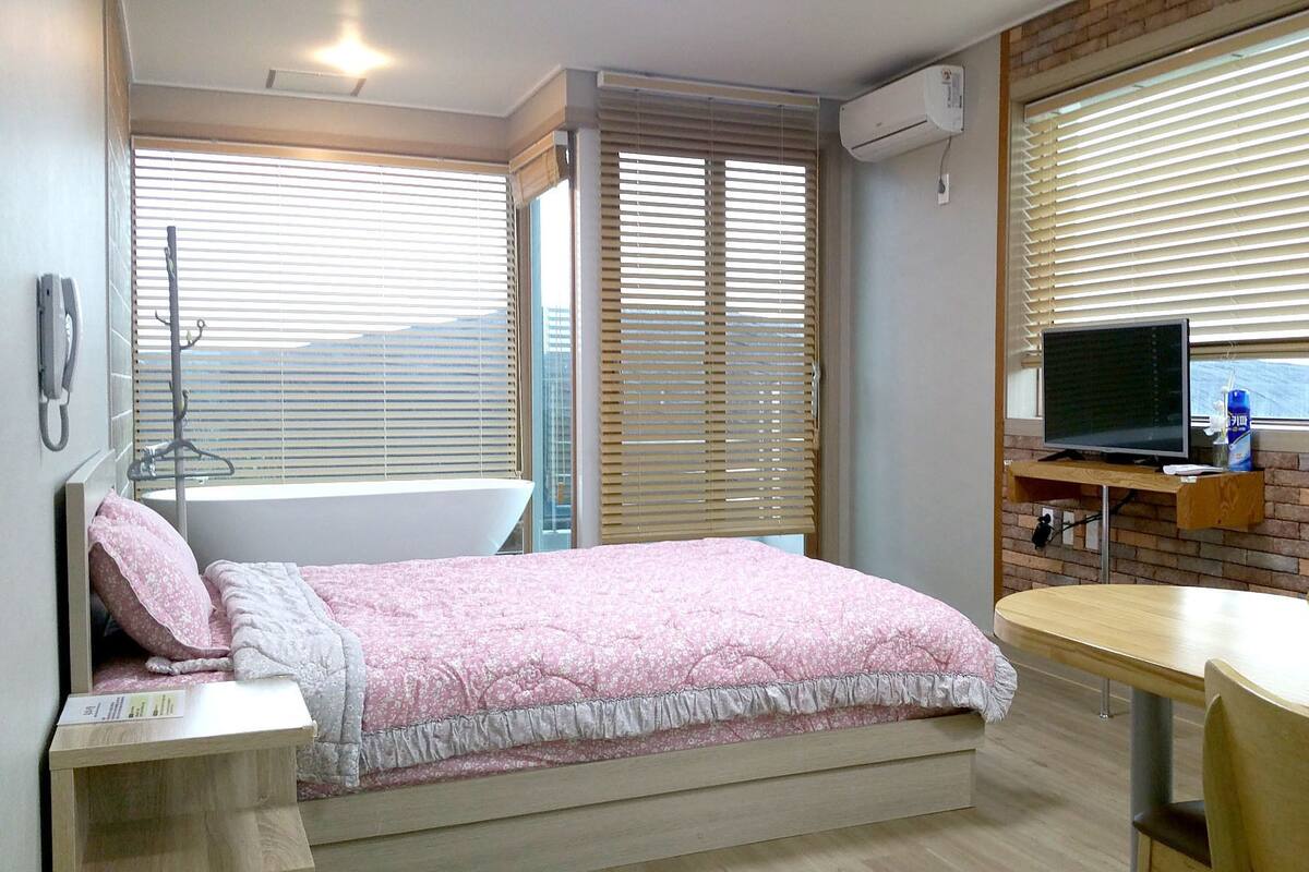 A bedroom with a bed, a desk, a chair, a TV, and a window with blinds.