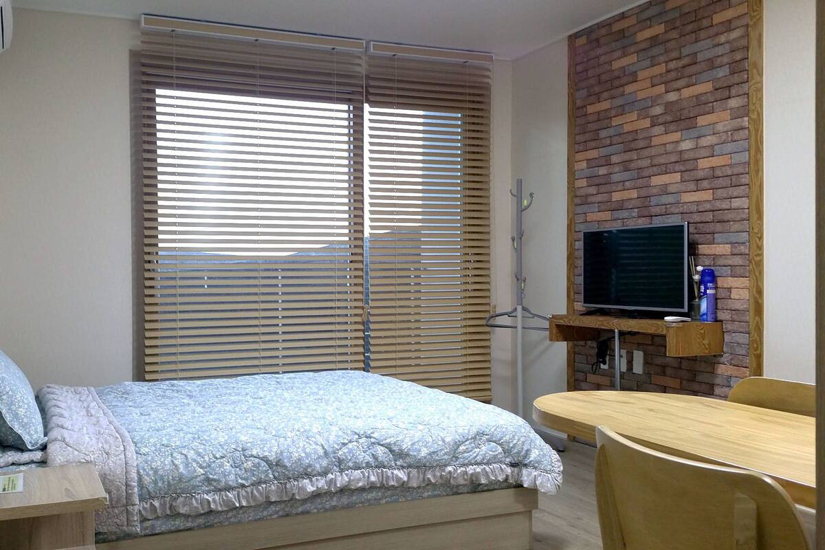 A bedroom with a bed, a desk, a television on a brick wall, and a window with blinds.
