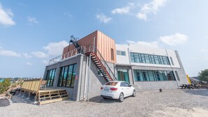 A modern building with a car parked in front, a staircase leading to a deck, and a picnic table nearby. - Pension Jung Won (Seogwipo)