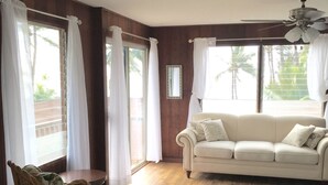 Un salon avec un canapé beige, un fauteuil en osier, un tapis à motifs et de grandes fenêtres ornées de rideaux blancs.