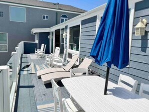 Een overdekt terras met witte tafels en stoelen, een blauwe parasol, en een huis met grijze gevelbekleding en een donker dak.