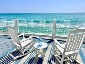 A wooden deck with white rocking chairs and a small table, overlooking a turquoise ocean. - Beachfront Fun, Classic Beach House w/ Fire Ring (Oceanside)