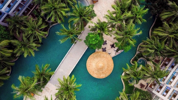 Luchtbeeld van een resort met een centraal gelegen zwembad, omringd door palmbomen, een pad en een rieten parasol.