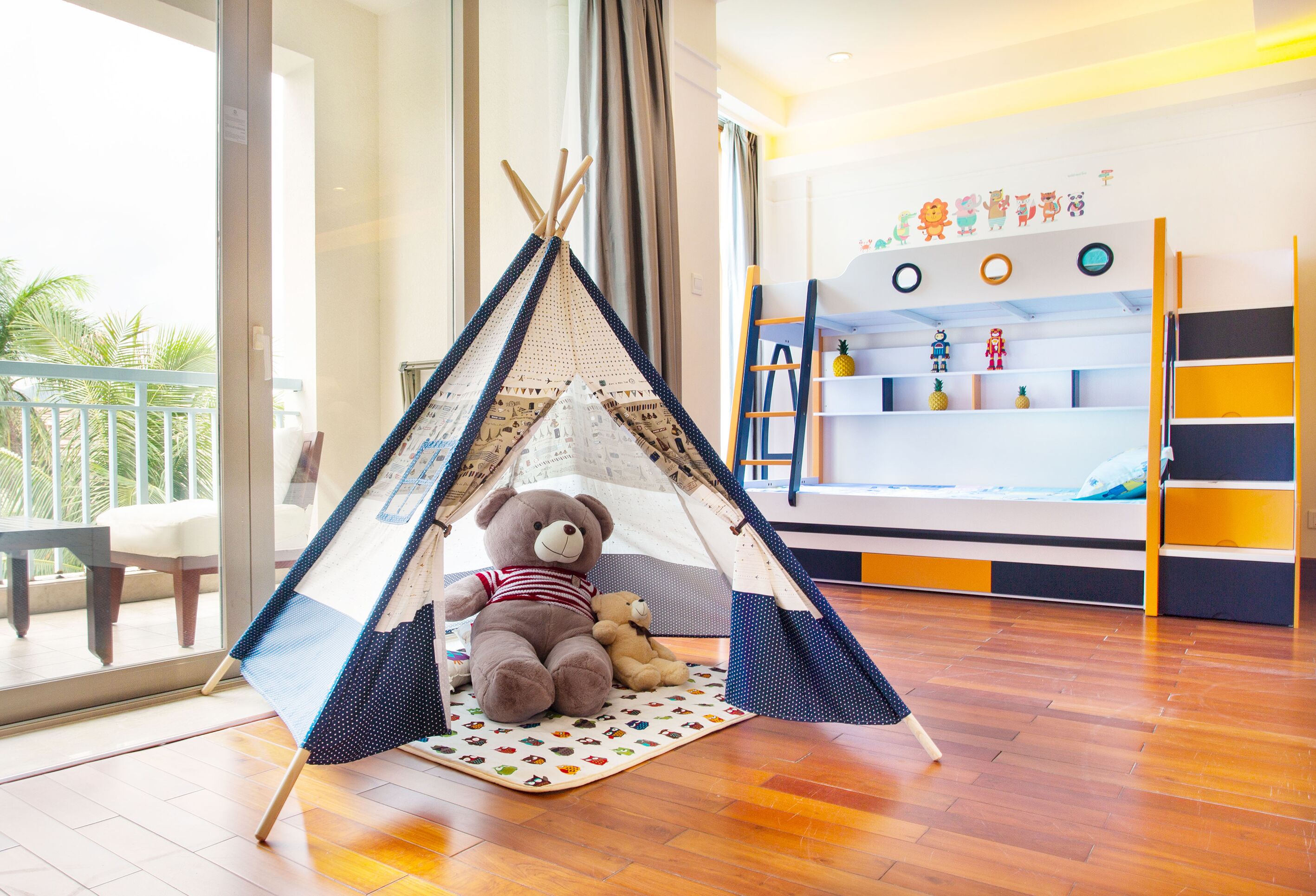 A room with a wooden floor, a tent-like play area with a teddy bear, a bookshelf with children's toys, and a balcony with a view of palm trees.