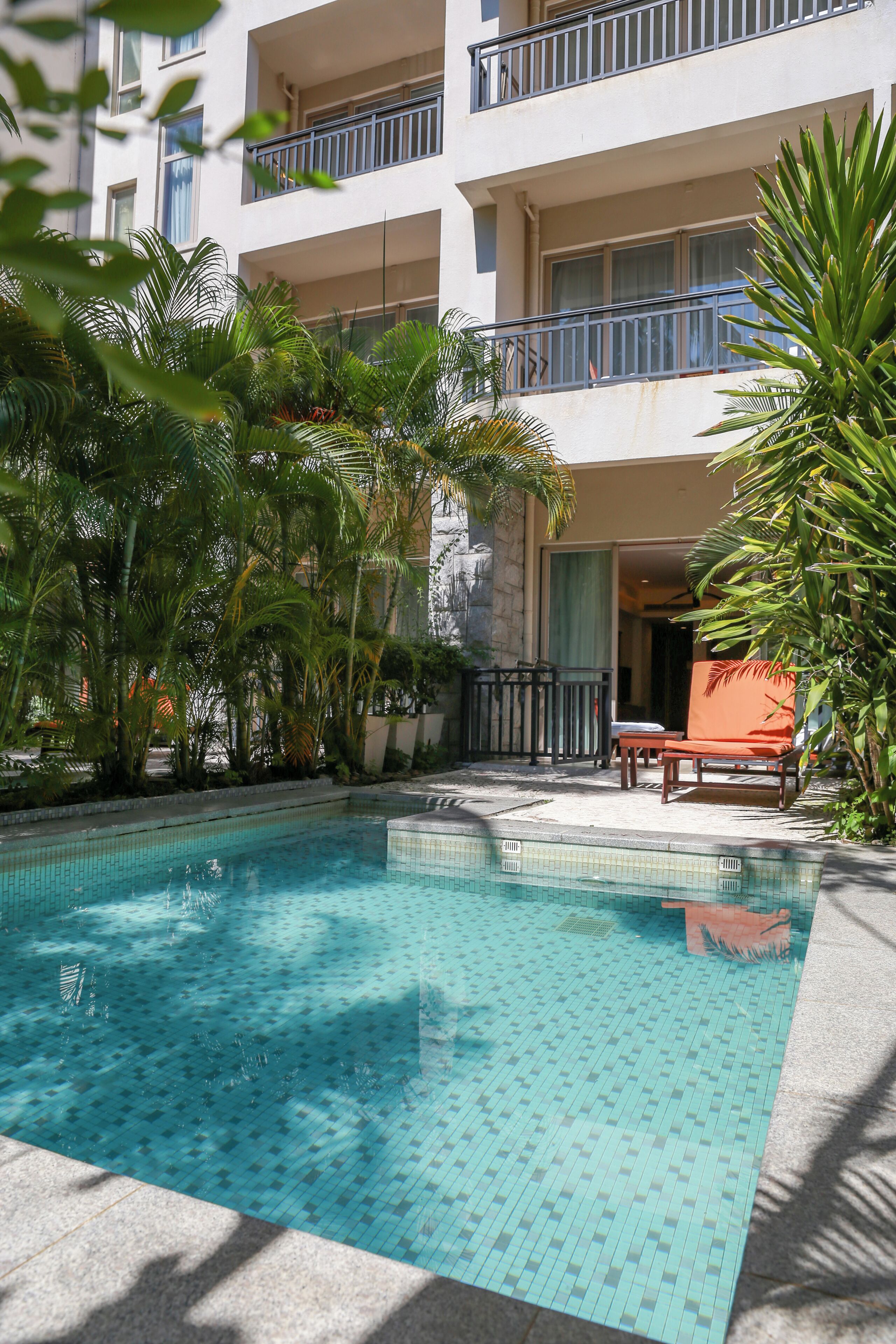 Una piscina con bordes de azulejos, rodeada de abundante vegetación y un edificio con balcones.