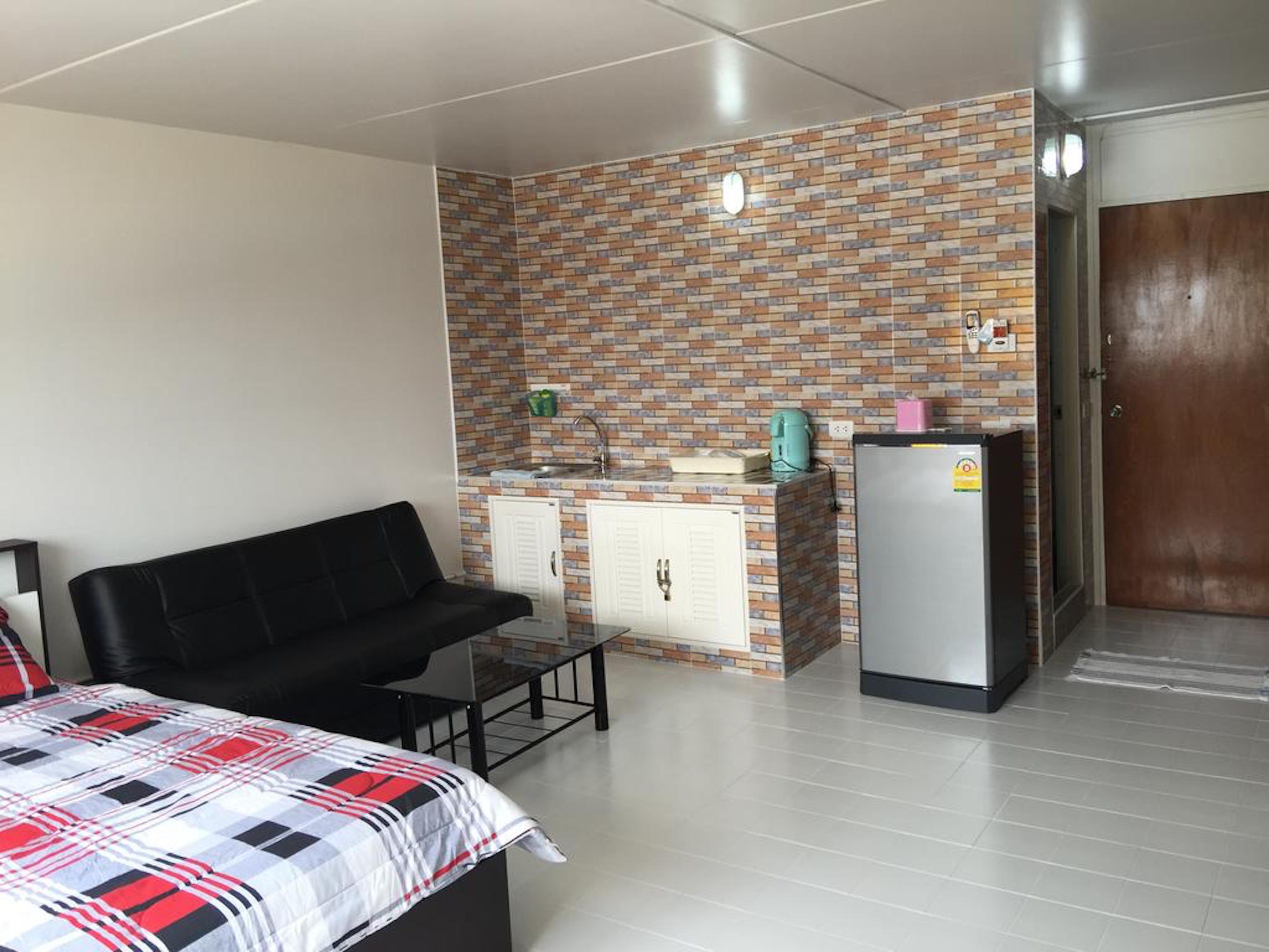 A compact room with a bed, a black sofa, a small table, a kitchenette with a fridge, and a wooden door.