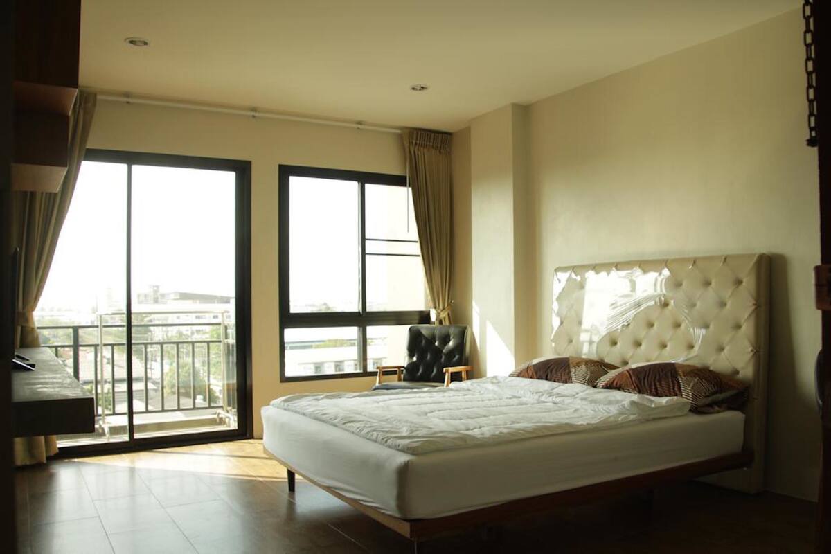 A bedroom with a large bed, a chair, a desk, and a view of the outside through a large window.