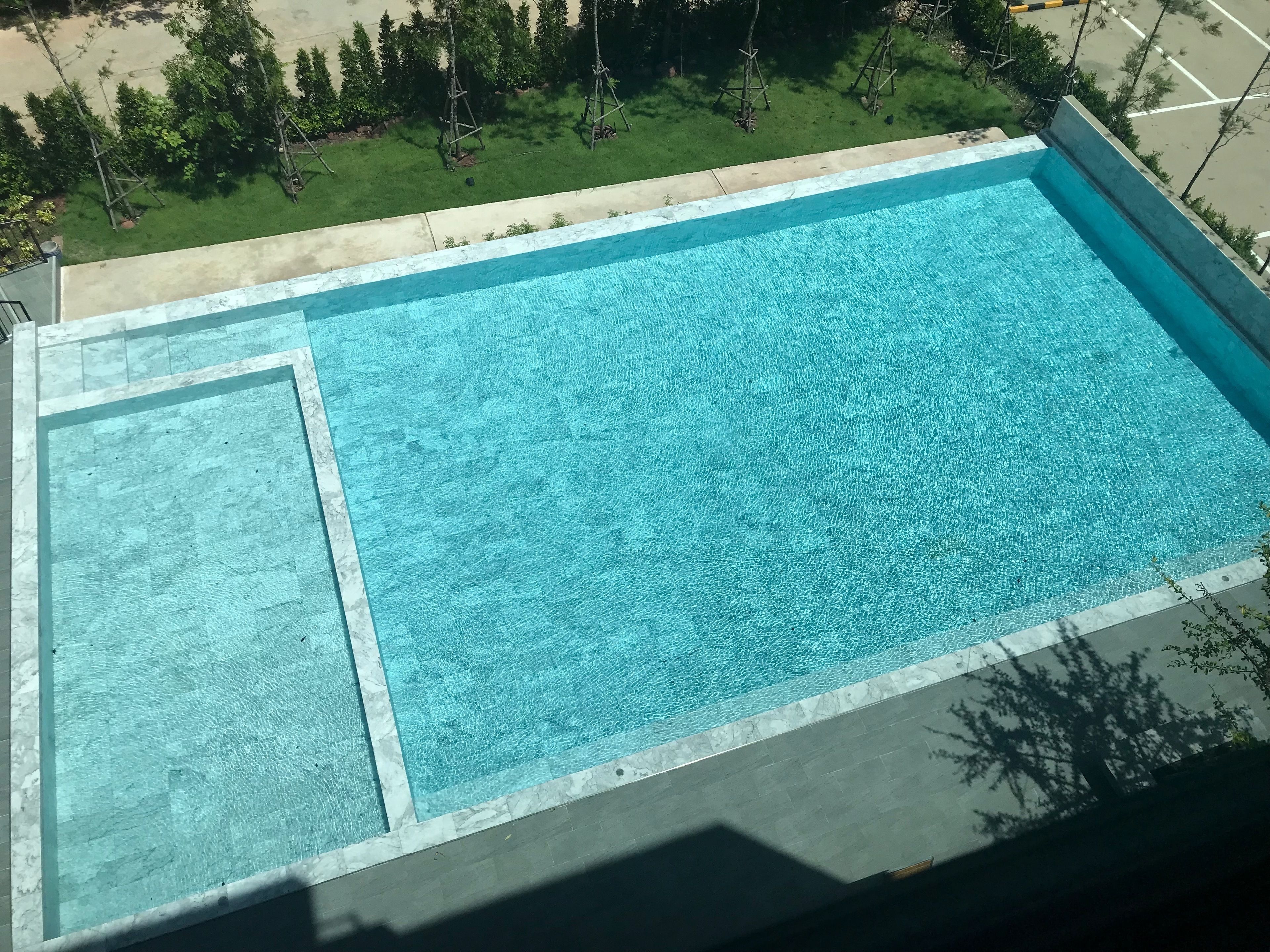 A rectangular swimming pool with a clear blue water surface, surrounded by a tiled deck and a grassy area with trees.