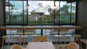 An indoor seating area with tables and stools, large windows with a view of buildings and trees, and hanging pendant lights. - The Residence Thepkanjana (Krathum Baen)