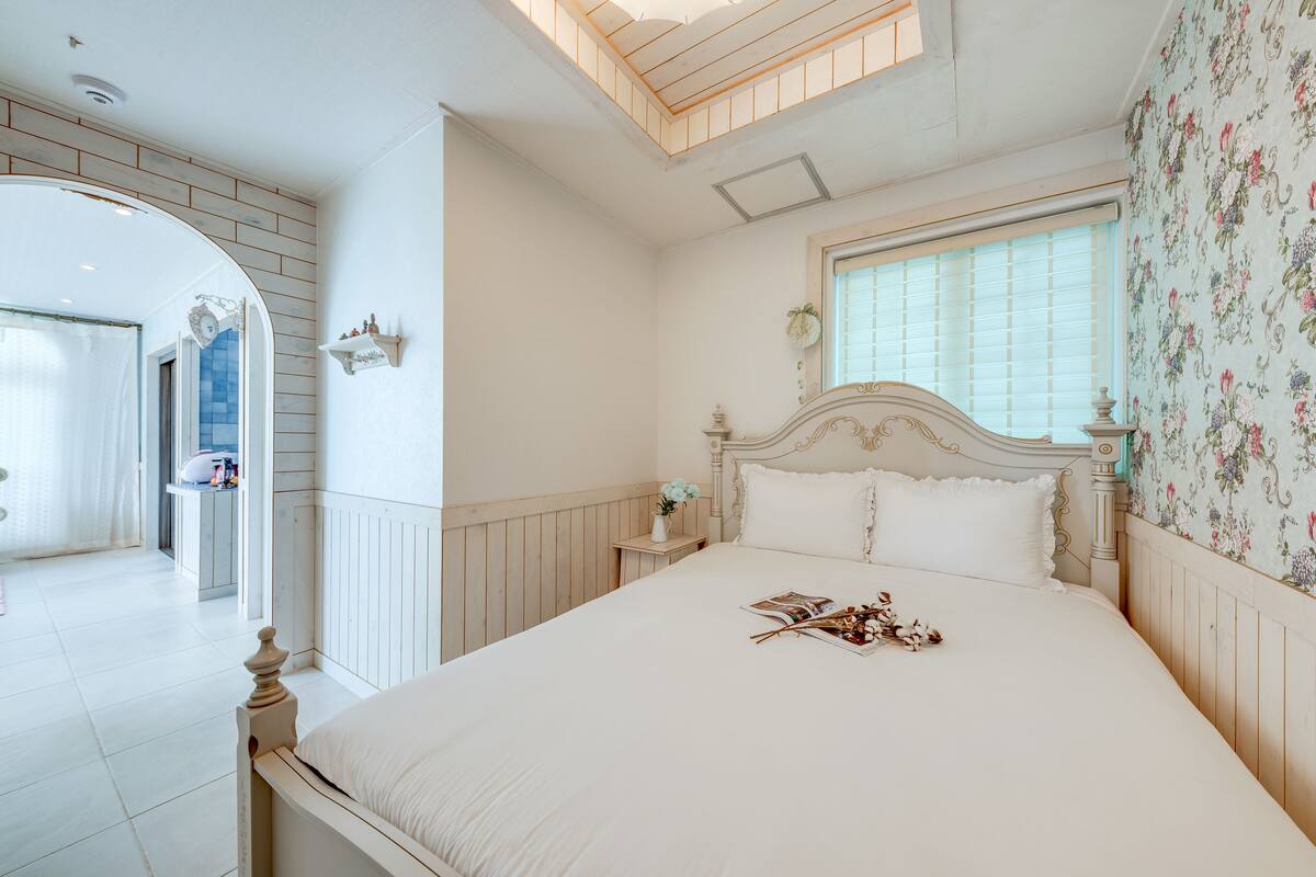 Une chambre avec un grand lit, une table de chevet, une fenêtre avec des stores et un papier peint à motifs floraux.