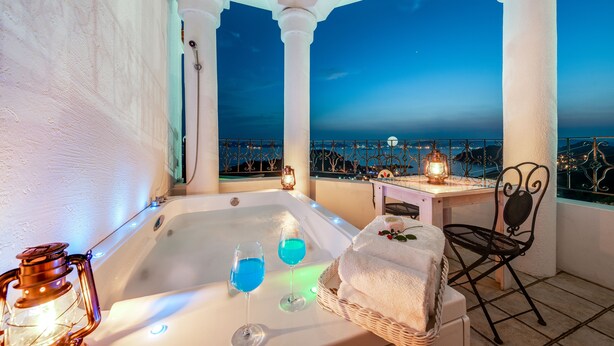 Un balcon avec un jacuzzi, deux verres contenant un liquide bleu, un panier en osier, une table avec une bougie et une chaise.