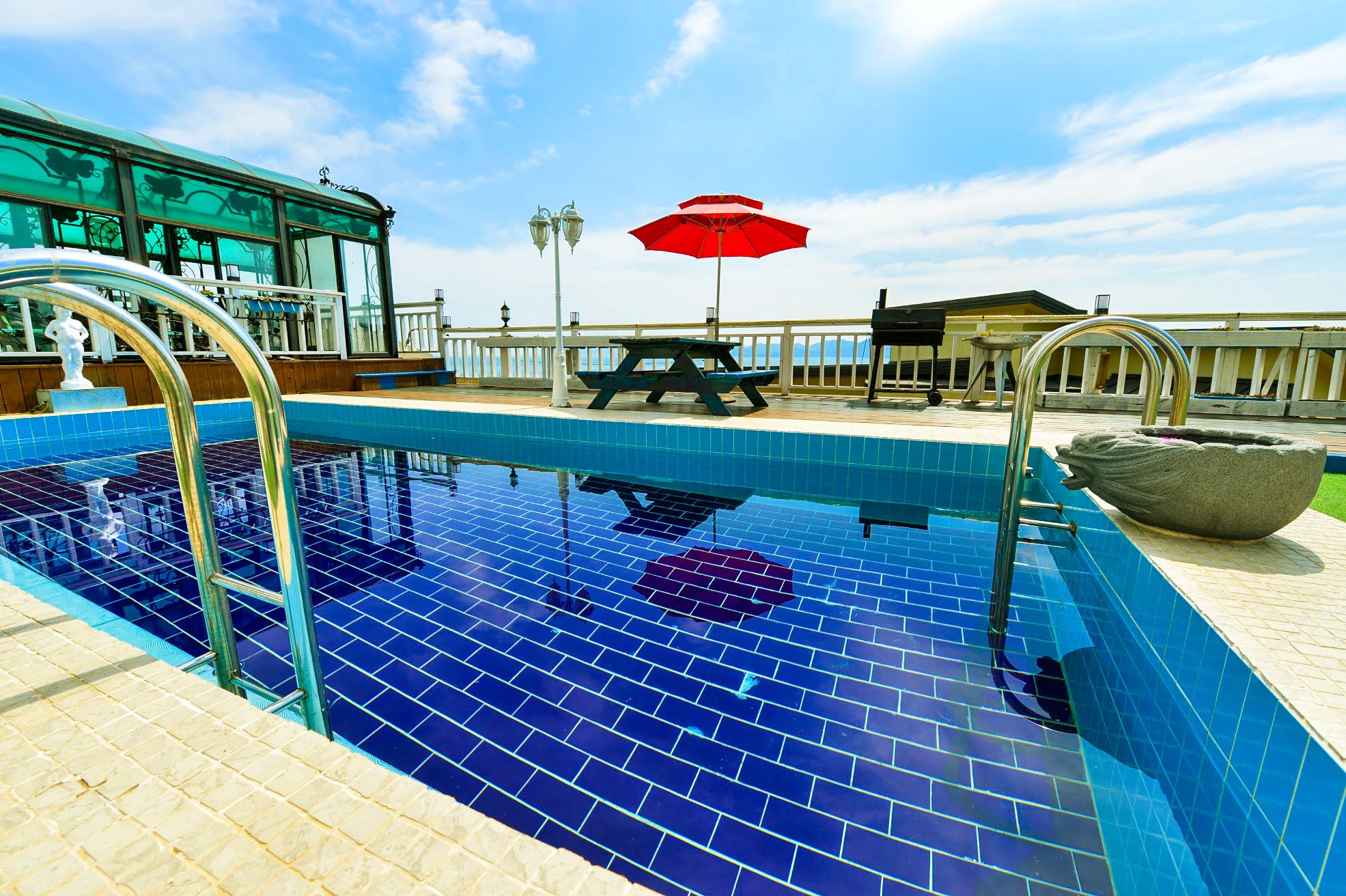 A swimming pool with a blue tiled deck, a red umbrella, and a stone sculpture.
