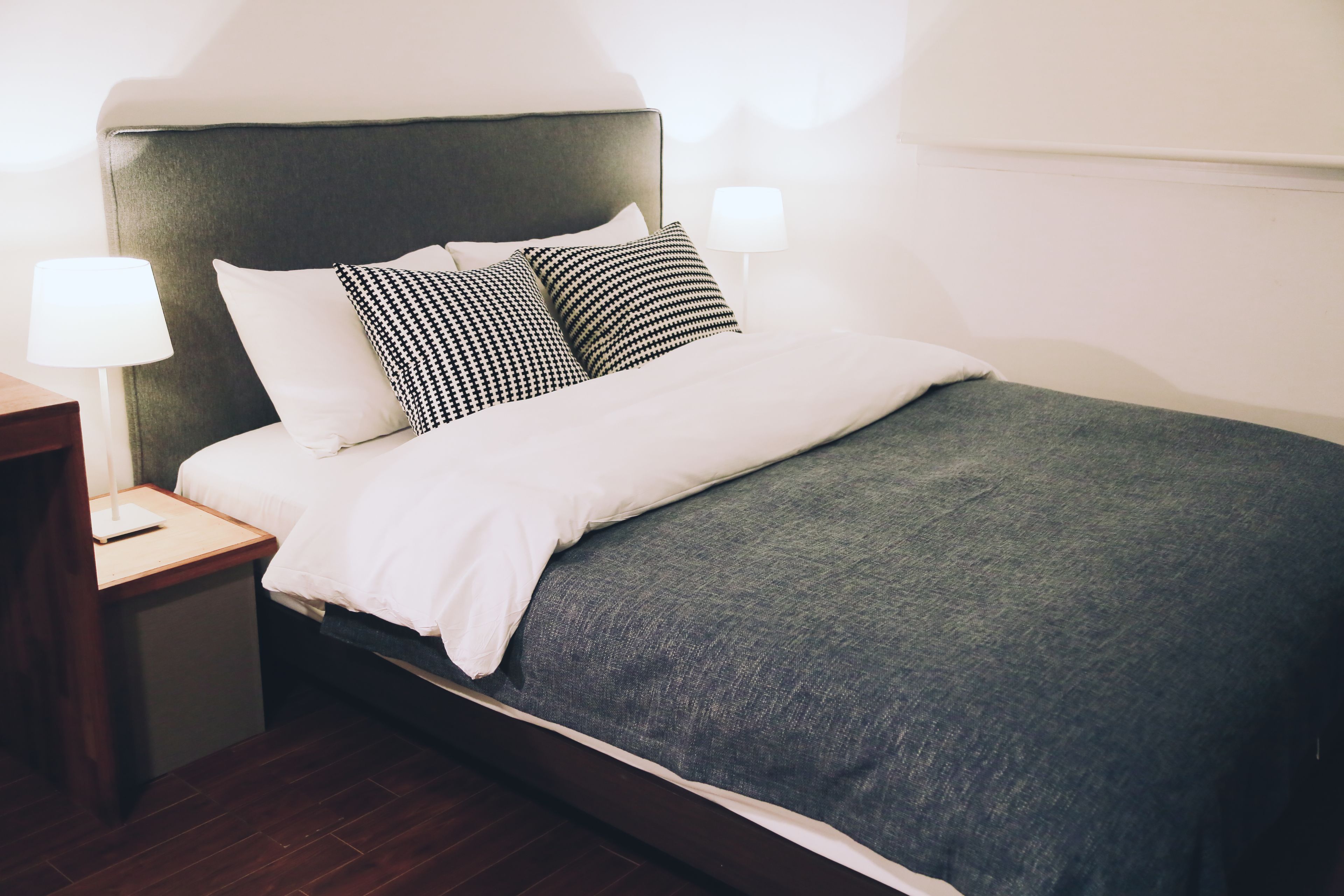 A neatly made bed with a grey headboard, white bedding, and two black-and-white patterned pillows. There is a bedside table with a lamp and a small electronic device.
