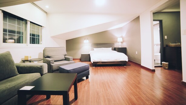 A modern hotel room with a large bed, a sofa, a coffee table, and a desk. There is a window with blinds and a door leading to the bathroom. - GONGGAN Residence Hotel (Geoje)