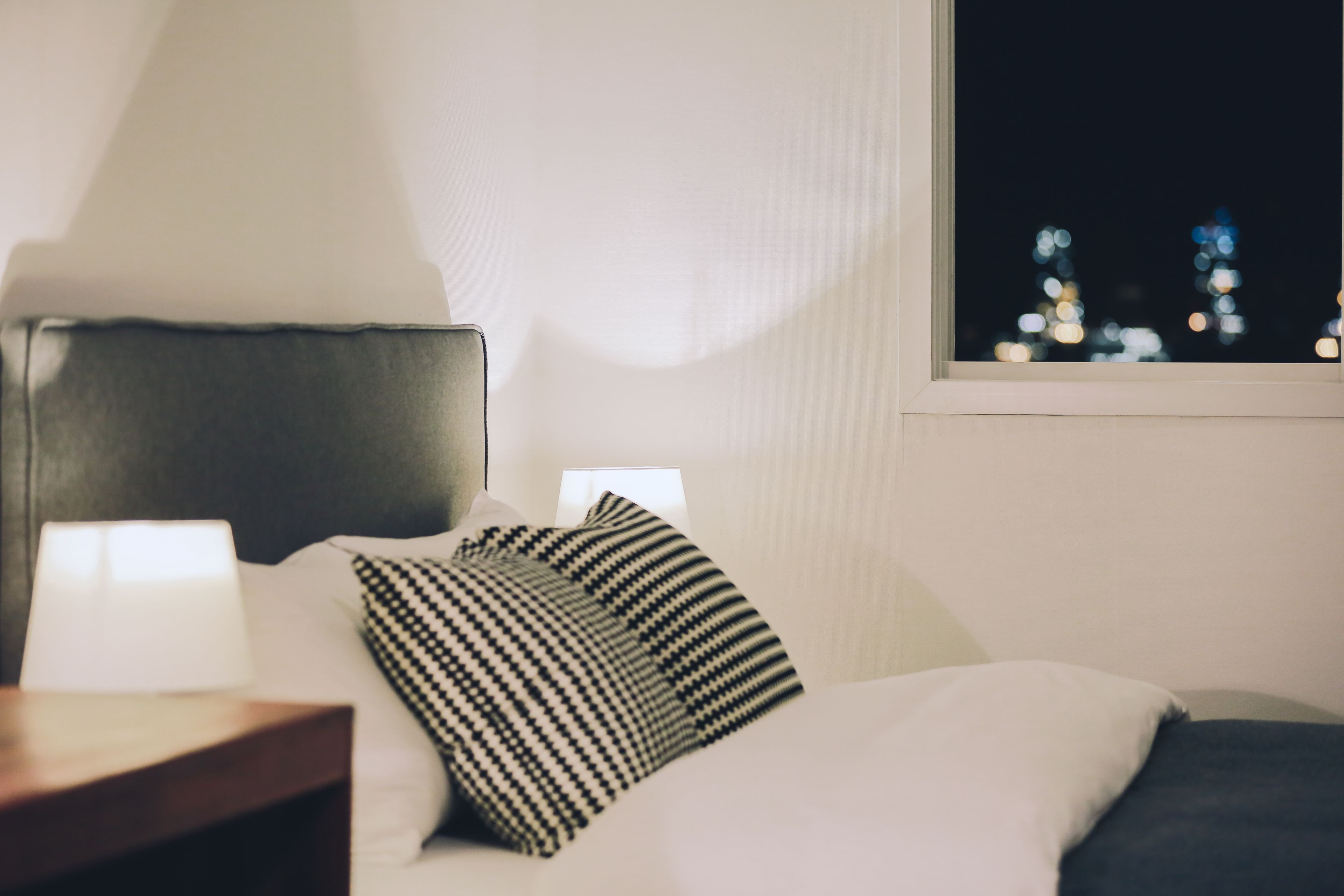 A neatly made bed with a striped pillow, a bedside lamp, and a view of city lights through a window.