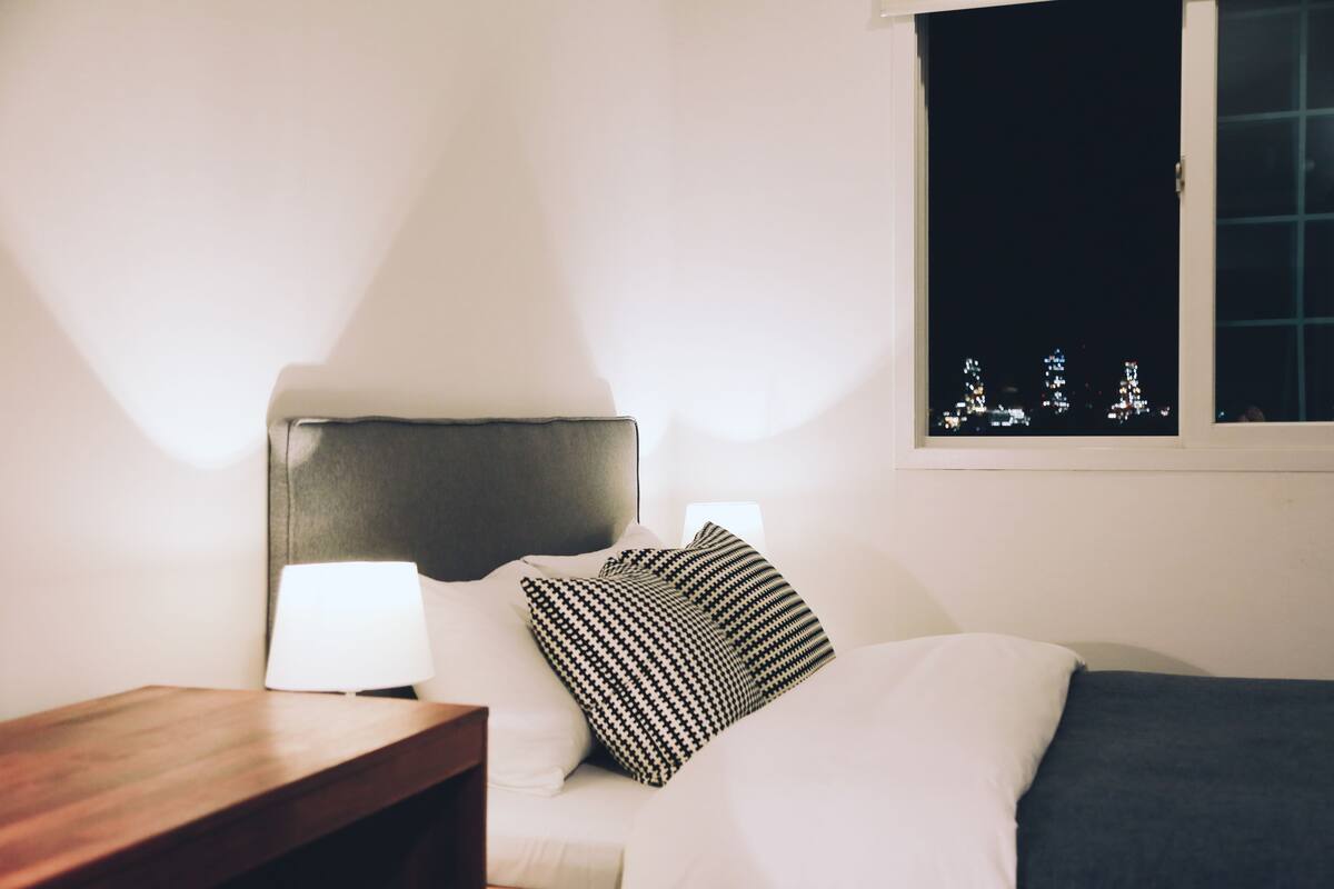Una cama bien hecha con cabecera, una lámpara de noche y una ventana con vistas a las luces de la ciudad.