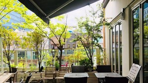 An outdoor patio area with wooden tables and chairs, surrounded by potted plants and trees. - GONGGAN Residence Hotel (Geoje)