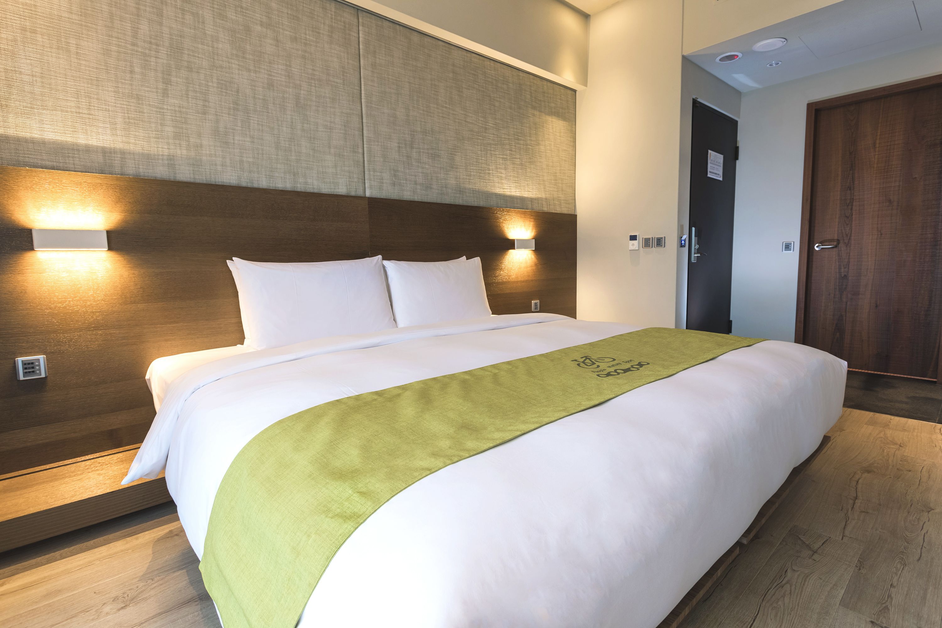 A neatly made bed with a green bedspread, two wall-mounted lights, and a wooden headboard.