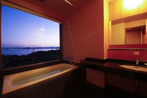 Ein modernes Badezimmer mit einer großen Badewanne, einer Dusche und einem Waschbecken. Es besitzt ein großes Fenster mit Blick auf Wasser und Hügel.
