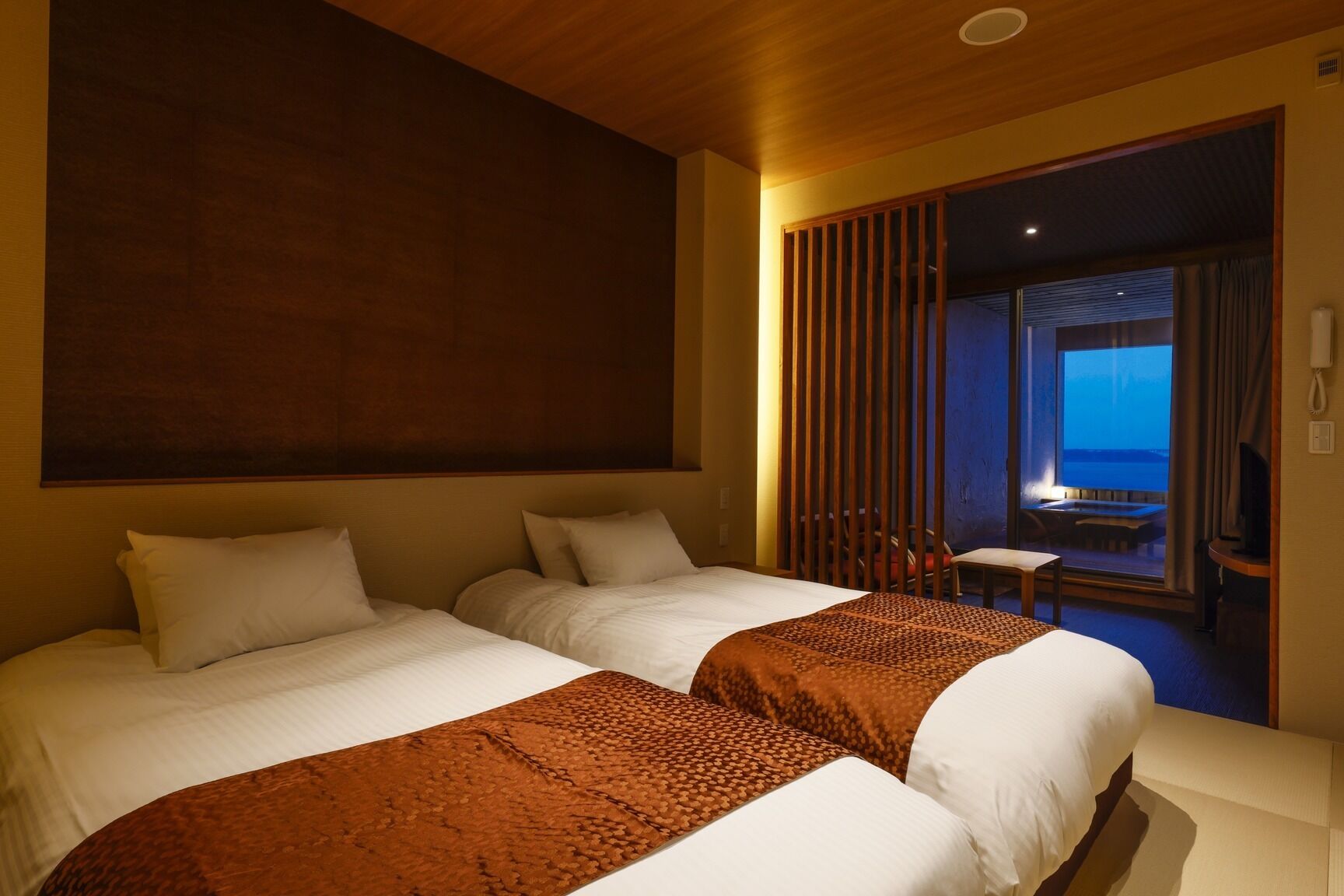 A hotel room with two beds, a view of the sea, a balcony with a table and chairs, and a wooden ceiling.