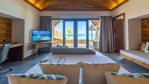 A bedroom with a large bed, a desk with a computer, a TV, and a view of the beach. - Fushifaru Maldives (Fushifaru)