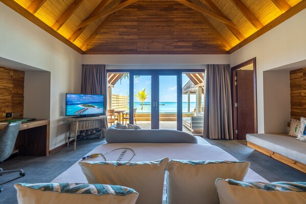 A bedroom with a large bed, a desk with a computer, a TV, and a view of the beach. - Fushifaru Maldives (Fushifaru)
