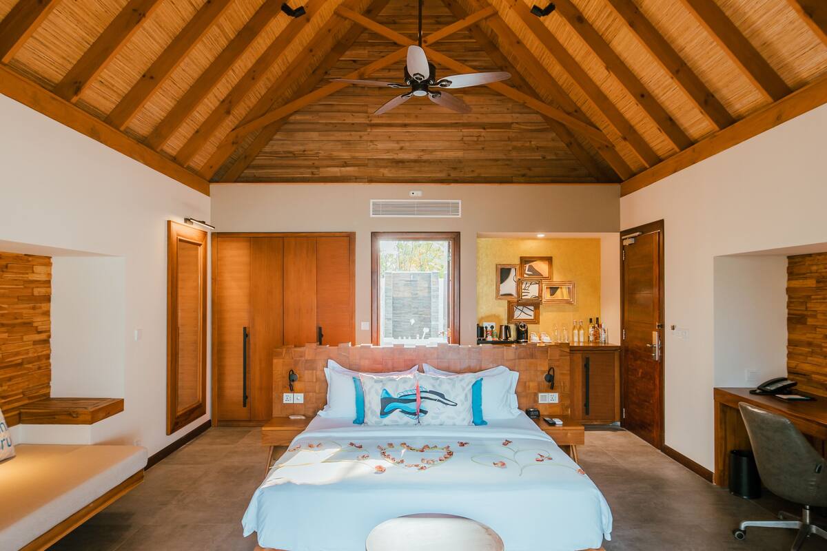 A spacious bedroom with a high ceiling, a bed with a dolphin-themed pillow, a round coffee table, and a desk with a chair.