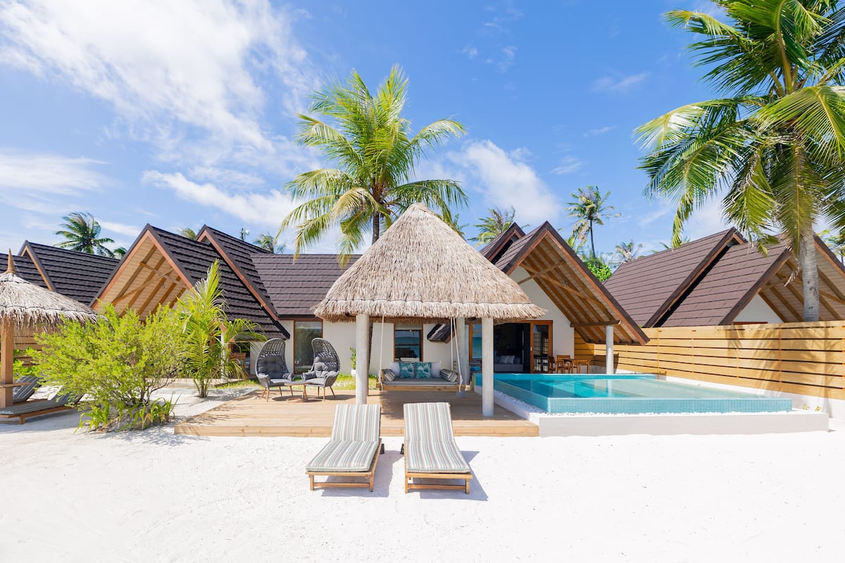 A resort with thatched-roof overwater bungalows, a swimming pool, and lounge chairs on a sandy beach.