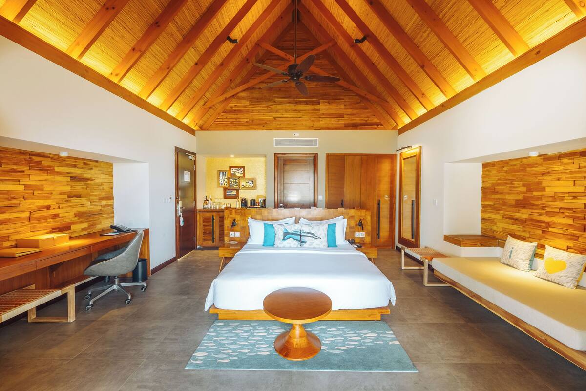 A spacious bedroom with a large bed, wooden ceiling beams, a desk, and a seating area.