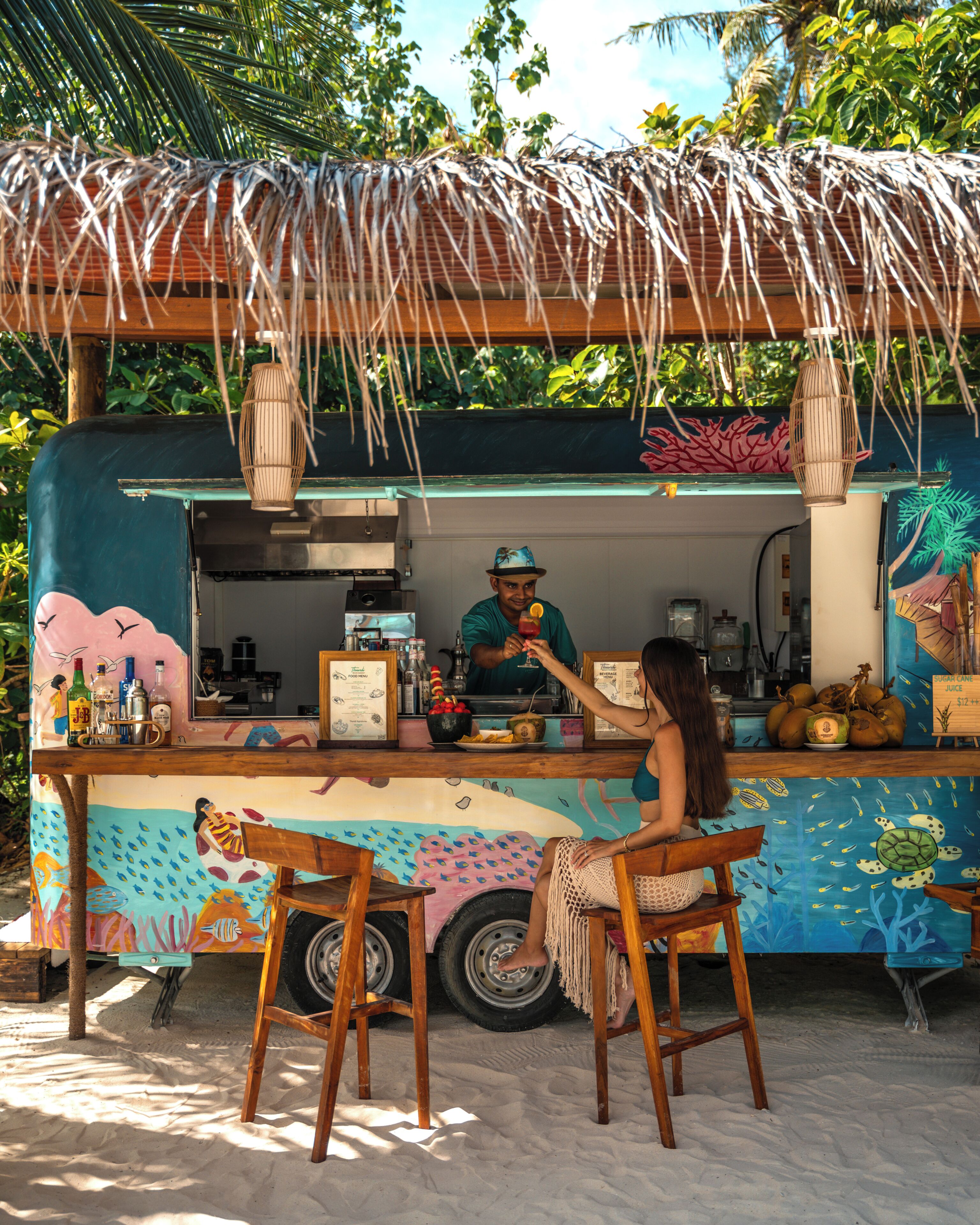 Un carrito de comida con un mural tropical, un cocinero preparando alimentos y dos sillas de madera.