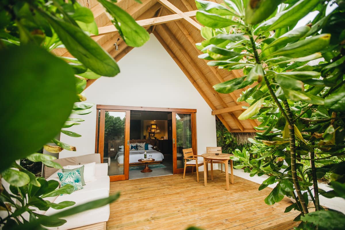 A wooden deck with a bed, a table, and chairs, surrounded by greenery.