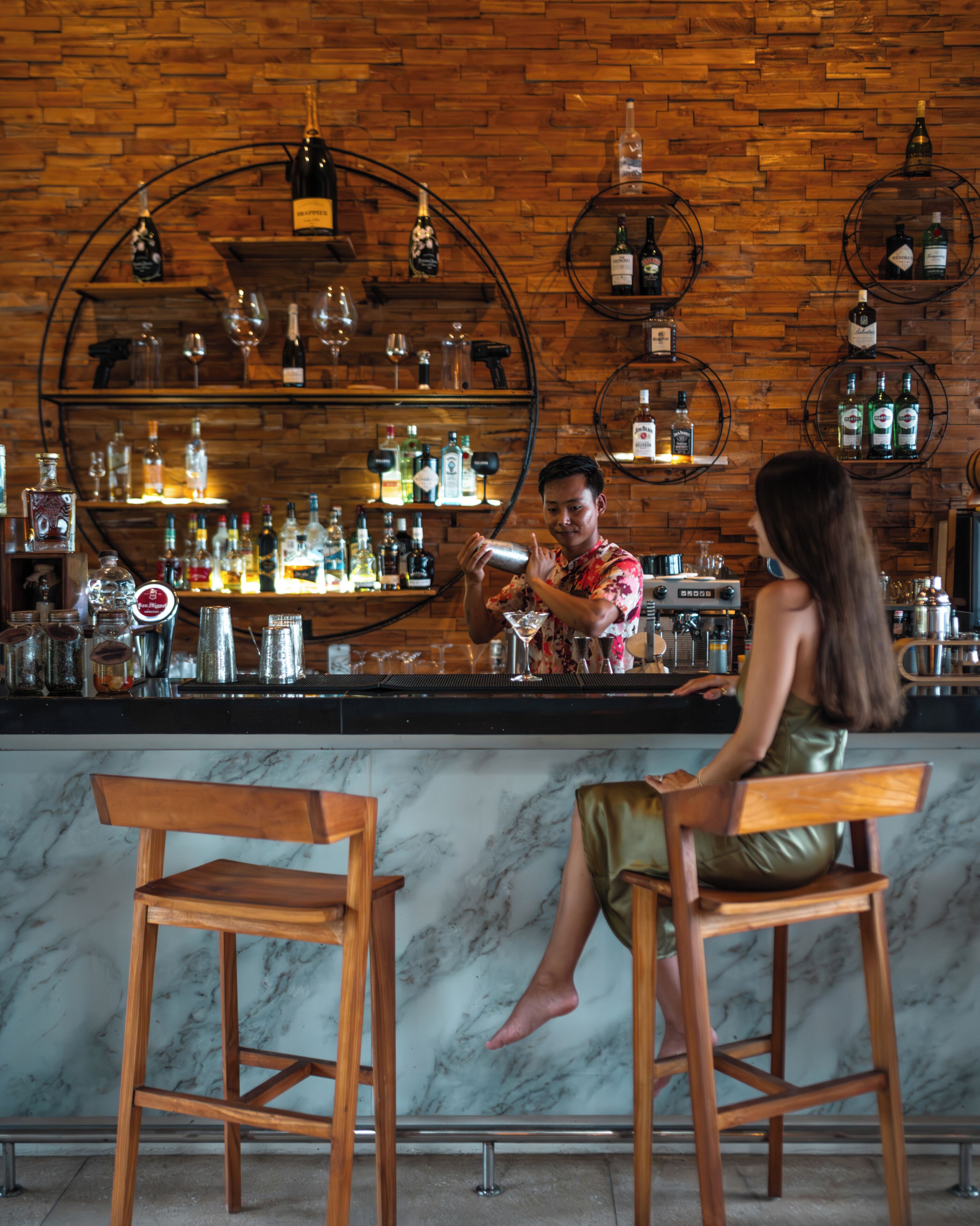 A bar with wooden shelves and a marble countertop, two wooden barstools, and a person preparing a drink.