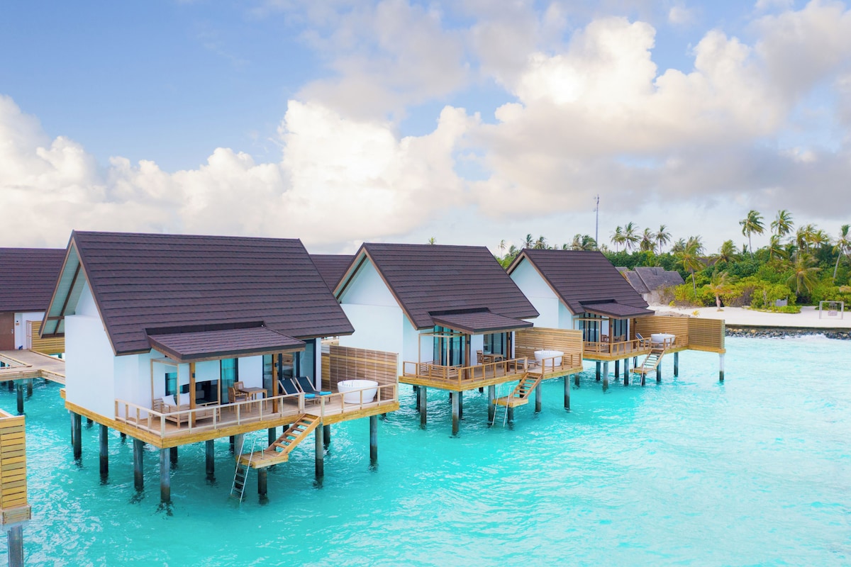 Overwater bungalows with wooden decks and glass walls, connected by walkways, set in clear turquoise waters.