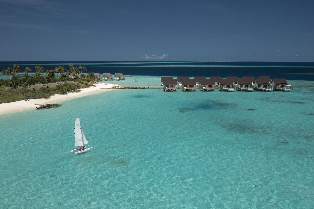 An overwater bungalow resort with clear turquoise waters, a sandy beach, and a small sailboat.