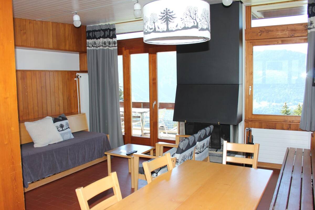 A cozy room with a bed, a table, chairs, a fireplace, and large windows offering a view.